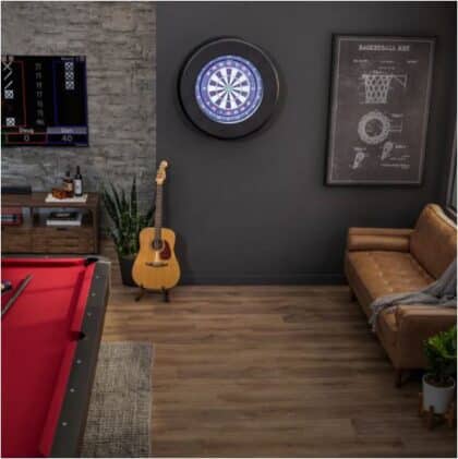 Game room with a lit dartboard on the dark wall, a guitar on a stand, a red-edged pool/billiards table, and a brown sofa with a throw blanket on the right.