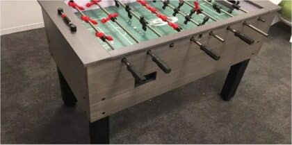 Foosball table with red and black players on a green field, metal sides and black legs in a room with a gray carpeted floor.