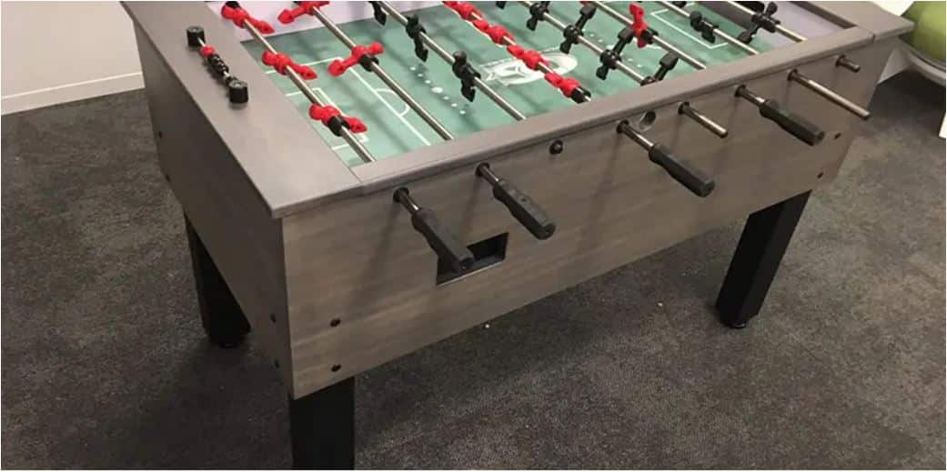 Foosball table with red and black players on a green field, metal sides and black legs in a room with a gray carpeted floor.