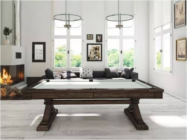 Dark wooden billiards table centered in a bright modern living room with a fireplace and a long sofa against a wall of windows.