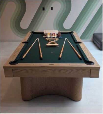 Wooden billiards/pool table with green felt, cues laid along the sides and a ball rack at the far end in a modern room.